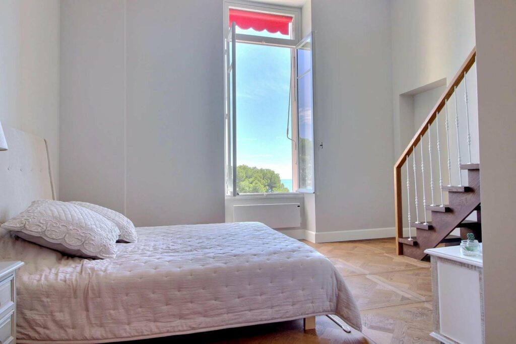 bedroom with light pink bedding next to large open windows with sea view